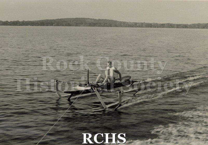 Black and white photograph of a hydrofoil boat with a young man in it as it moves through the water.