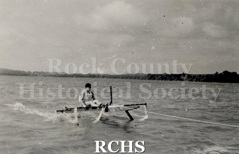 Black and white photograph of a hydrofoil boat with a young man sitting in it which is moving through the water. There is a shoreline in the background with trees.