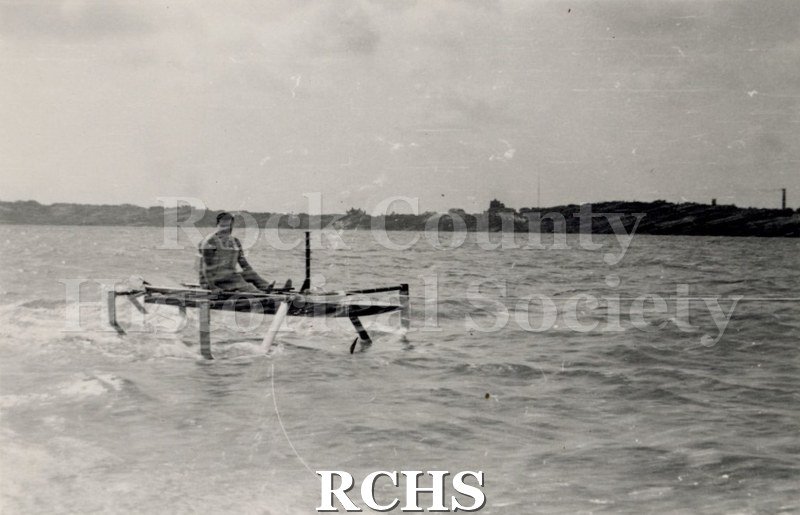 Black and white photograph of a hydrofoil boat moving through the water with a young man in it. There is a shoreline in the background with trees.