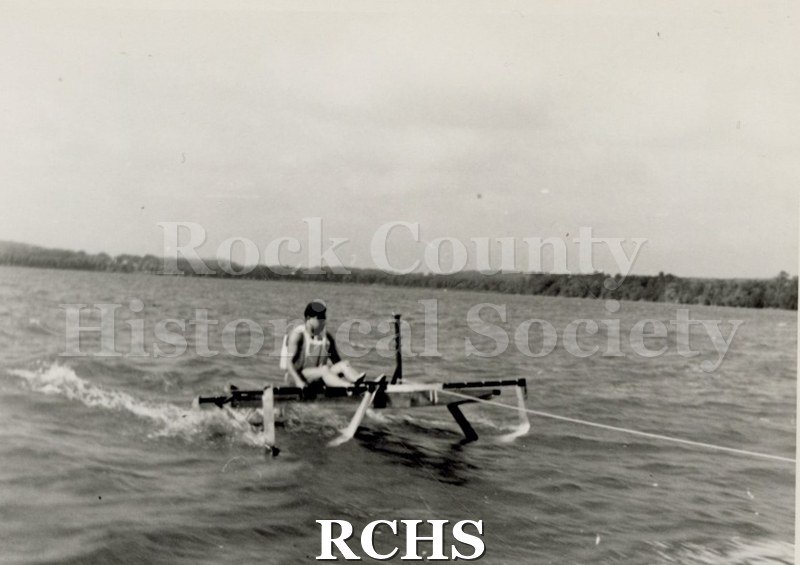 Black and white photograph of a hydrofoil boat moving through the water with young man sitting in it. There is a shoreline in the background with trees.