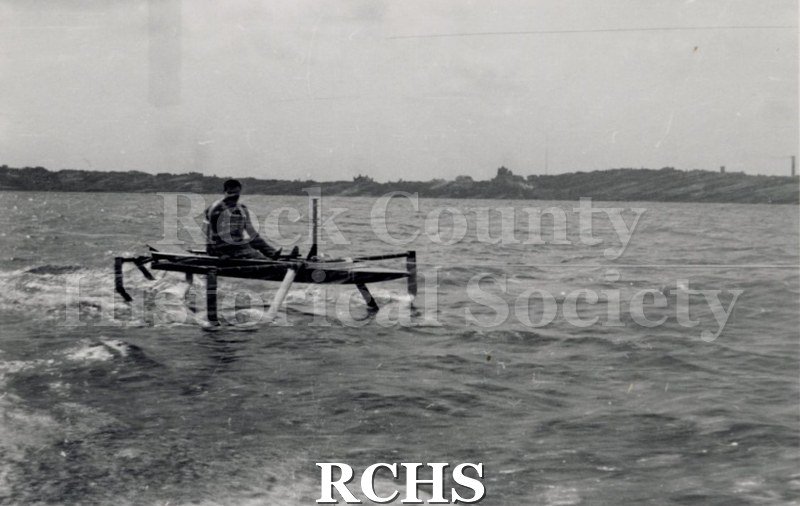 Black and white photograph of a hydrofoil boat moving through the water with a young man sitting in it. There is a shoreline in the background with trees.