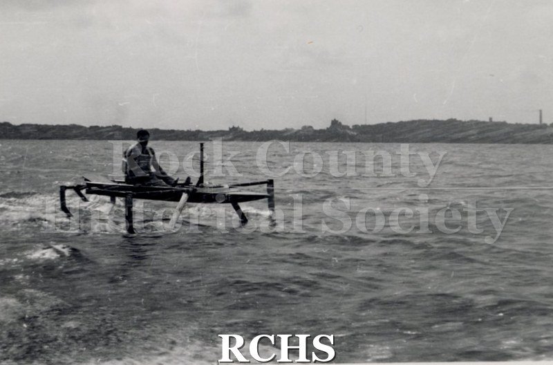 Black and white photograph of a hydrofoil boat moving through the water with a young man sitting in it. There is a shoreline in the background with trees.