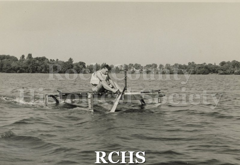 Black and white photograph of a hydrofoil boat moving through the water with a young man sitting in it. There is a shoreline in the background with trees.