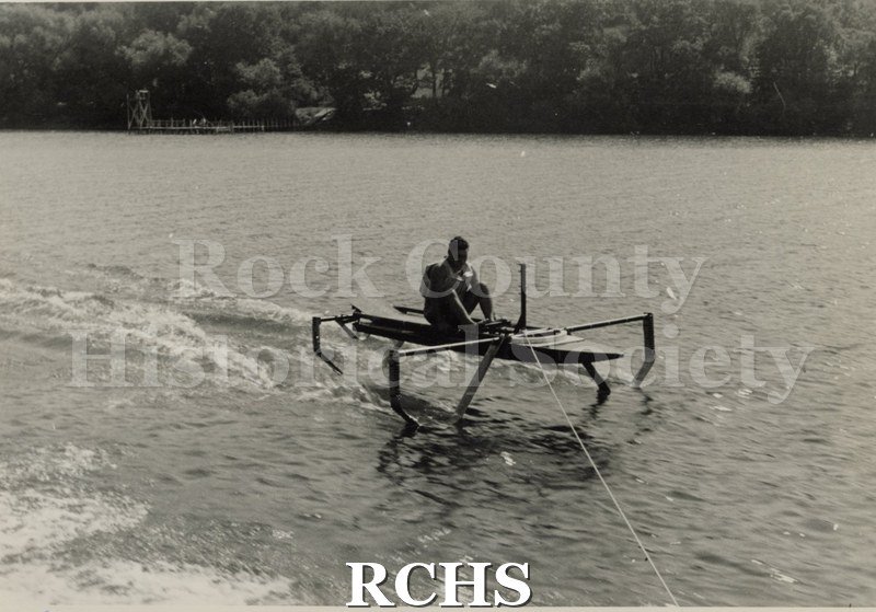 Black and white photograph of a hydrofoil boat moving through the water with a young man sitting in it. There is a shoreline in the background with trees.