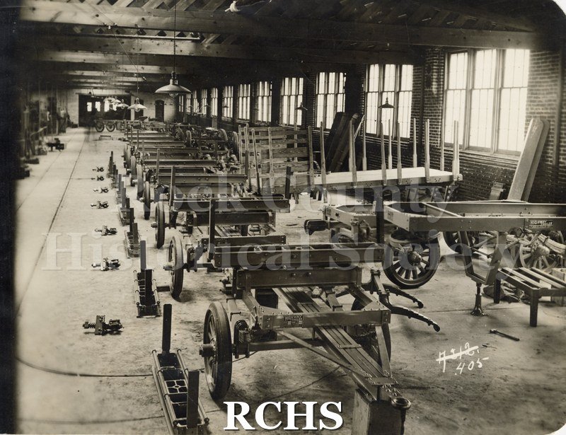 Black and white photograph of pole trailers and semi's from Highway Trailer Co., Edgerton, WI. Shows a factory with trailers lined up.