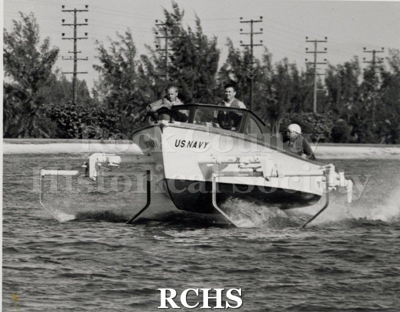 Black and white photograph of a US Navy boat on the water with three men aboard. On front of craft is: "USNAVY".