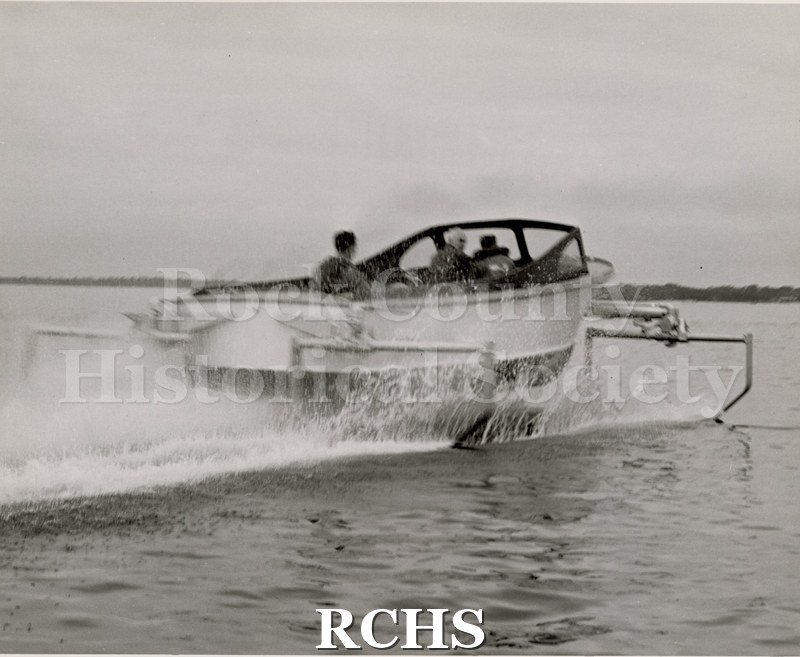 Black and white photograph of a Naval boat in the water with two men on board. Men are barely visible due to the water splashing up.