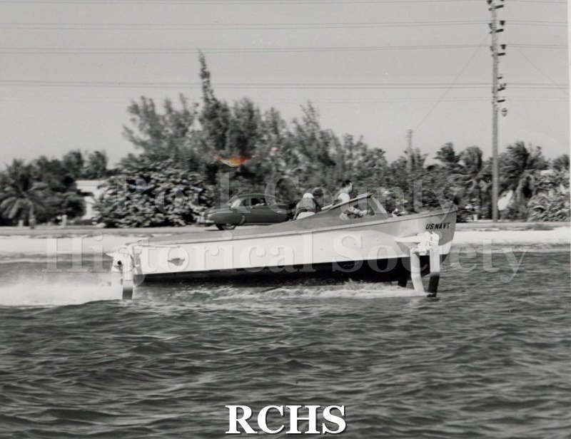 Black and white photograph of "High Pockets" with three men aboard. On front of craft is: "U.S.NAVY".