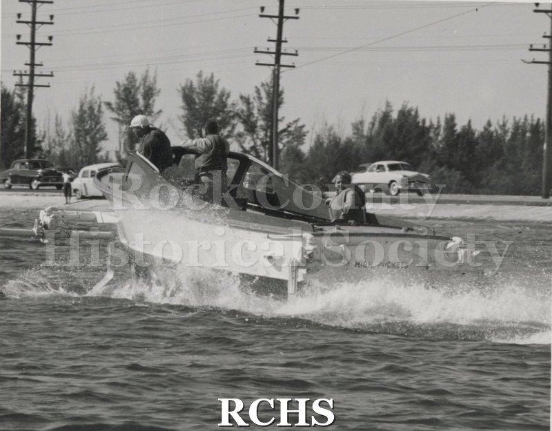 Black and white photograph of "High Pockets" with three men aboard. On back is the name: "High Pockets". Boat appears to be heading for a landing.