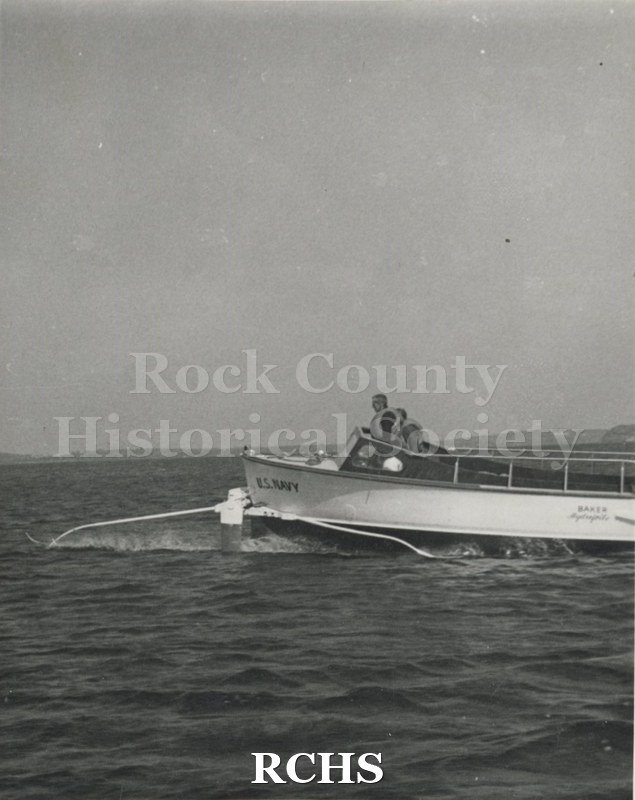 Black and white photograph of "High Pockets" in the water with two men in it. The name: "U.S.Navy" is visible. Also "Baker ____" is painted on the side of the boat.
