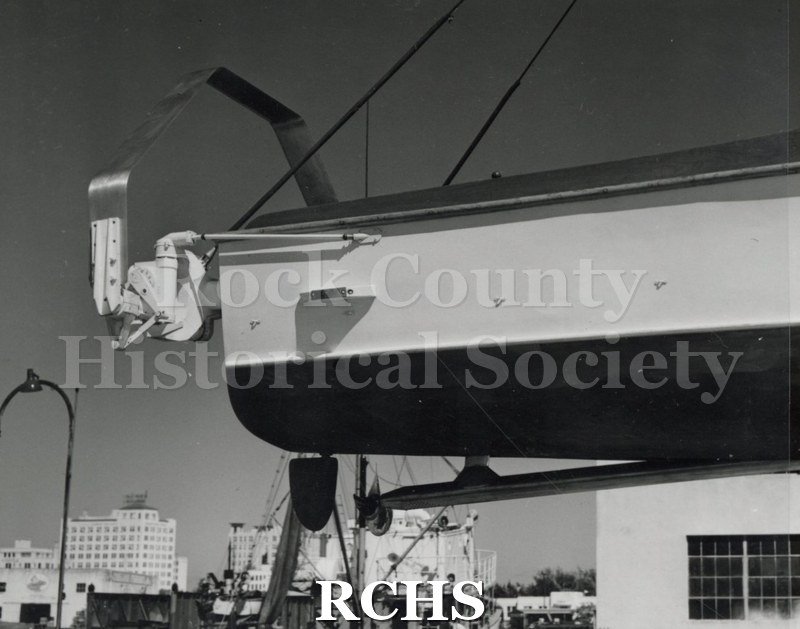 Black and white photograph of "High Pockets"(?) on a hoist above the water. Propellor is visible on the left side.