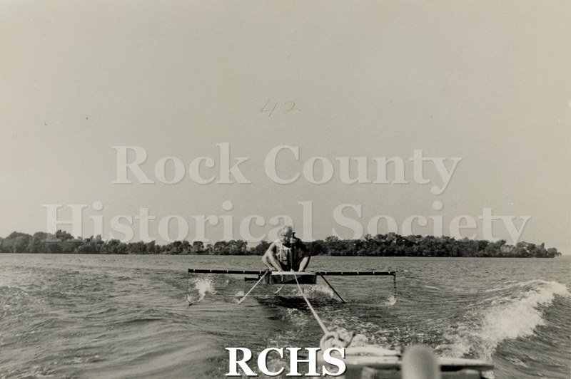Black and white photograph of a hydrofoil boat moving through the water with a young man sitting in it. There is a shoreline in the background with trees.