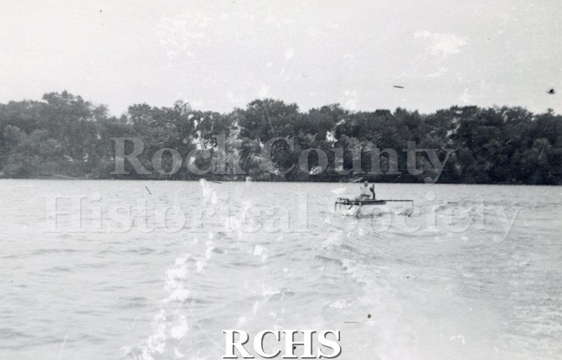 Black and white photograph of a hydrofoil boat moving through the water with a young man sitting in it.. There is a shoreline in the background with trees.