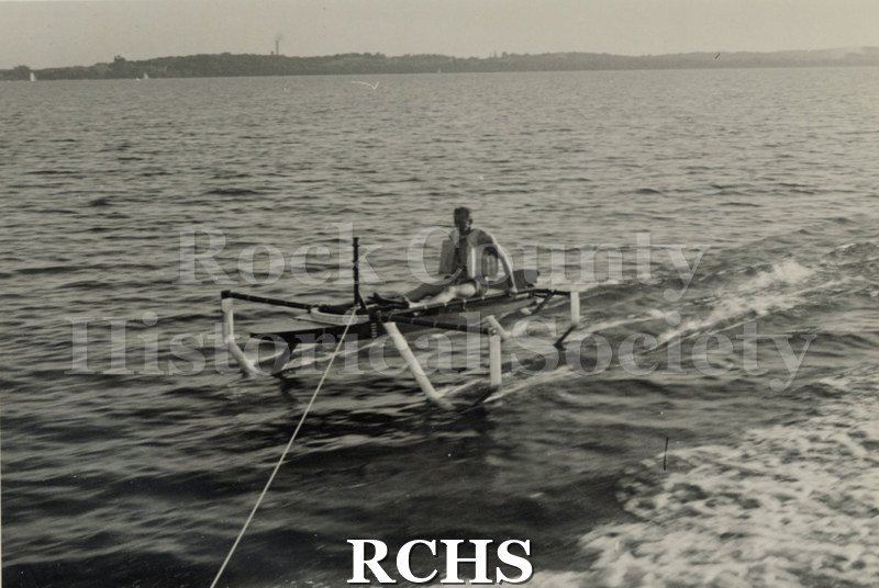 Black and white photograph of a hydrofoil boat stationary in the water with a young man sitting in it. There is a shoreline in the background with trees.