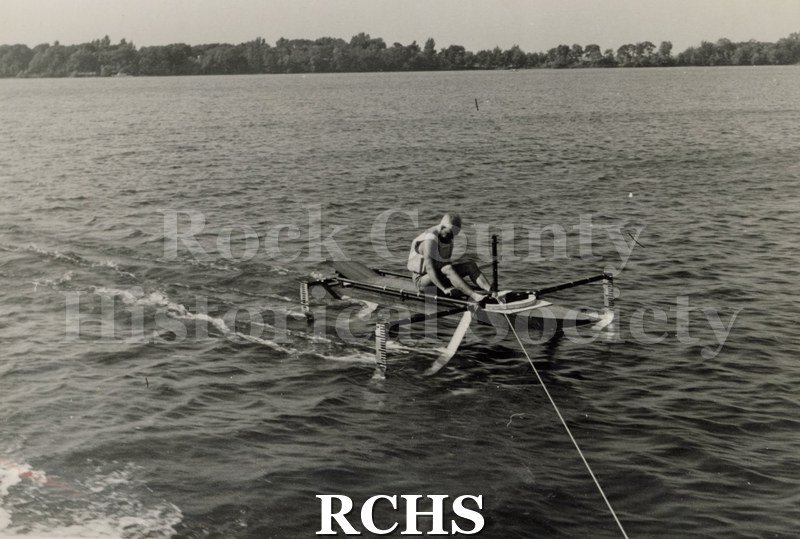 Black and white photograph of a hydrofoil boat stationary in the water with a young man sitting in it. There is a shoreline in the background with trees.