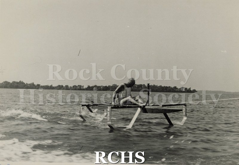 Black and white photograph of a hydrofoil boat stationary in the water with a person sitting in it. There is a shoreline in the background with trees.
