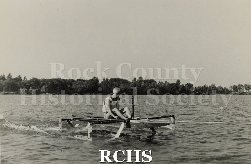 Black and white photograph of a hydrofoil boat stationary in the water with a person sitting in it. There is a shoreline in the background with trees.