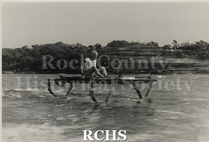 Black and white photograph of a hydrofoil boat stationary in the water with a man sitting in it. There is a shoreline in the background with trees.