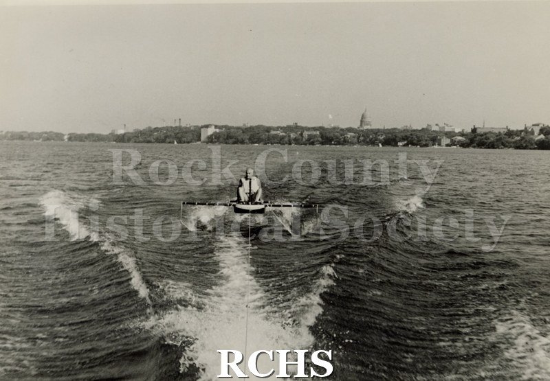 Black and white photograph of Foil Testing. There is a man sitting in a hydrofoil boat that is being towed. There is the shoreline of Madison in the background.