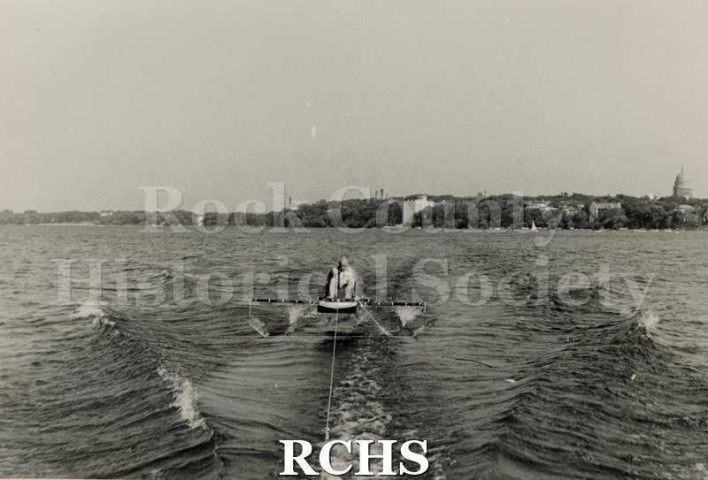Black and white photograph of Foil Testing. There is a man sitting in a hydrofoil boat that is being towed. There is the shoreline of Madison in the background.