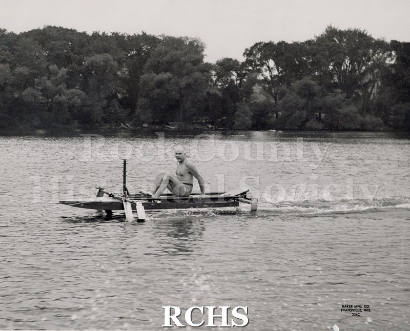 Black and white photograph of J. G. Baker in a boat which is stationary in the water. The man is looking at the camera. On bottom: Baker Mfg. Co. Evansville, Wis.