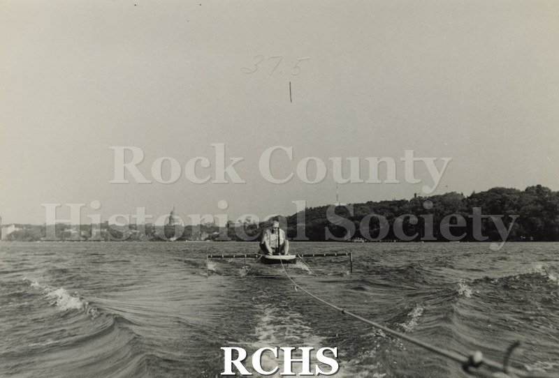 Black and white photograph of J.G. Baker sitting in a hydrofoil boat that is being towed. There is the shoreline of Madison in the background.