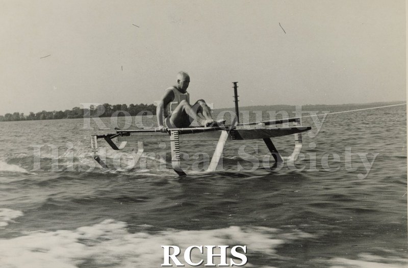 Black and white photograph of J.G. Baker sitting in a hydrofoil boat that is being towed. There is the shoreline of Madison in the background.