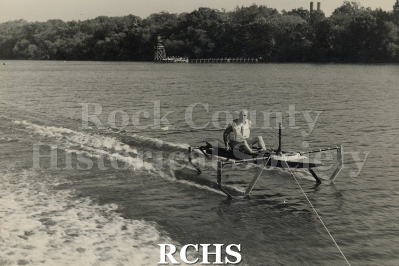 Black and white photograph of J. G. Baker sitting in a hydrofoil boat that is being towed. There is the shoreline of Madison in the background.