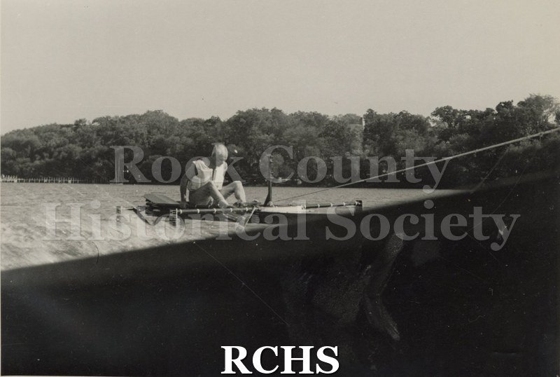 Black and white photograph of J. G. Baker sitting in a hydrofoil boat that is being towed. There is the shoreline of Madison in the background.
