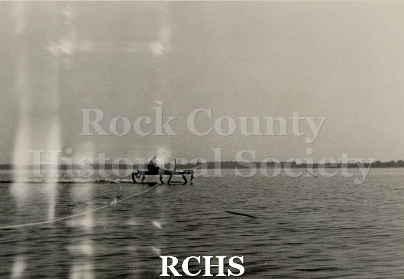 Black and white photograph of Warren Hull sitting on a hydrofoil in the water with a tow line visible.
