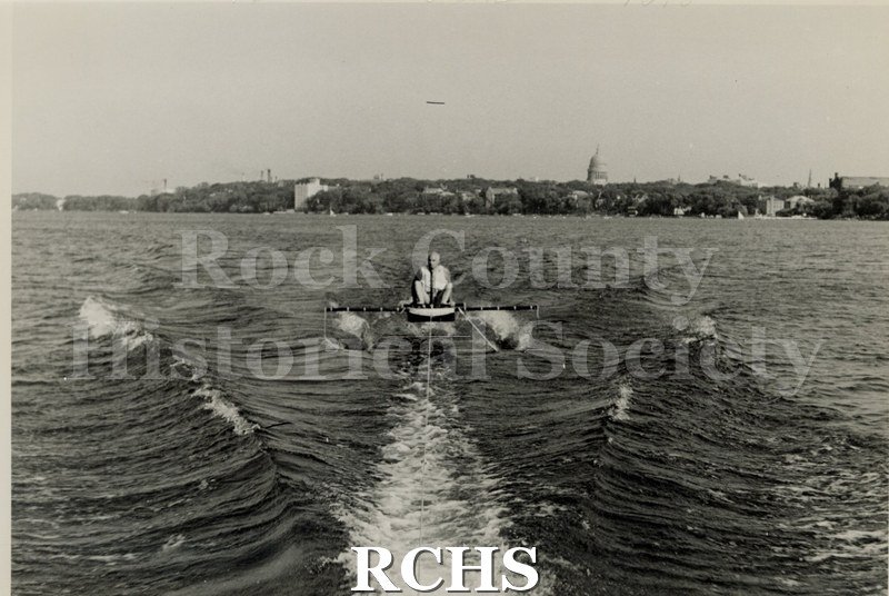 Black and white photograph of J. G. Baker sitting in a hydrofoil boat that is being towed. There is the shoreline of Madison in the background.