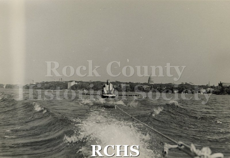Black and white photograph of J. G. Baker sitting in a hydrofoil boat that is being towed. There is the shoreline of Madison in the background.