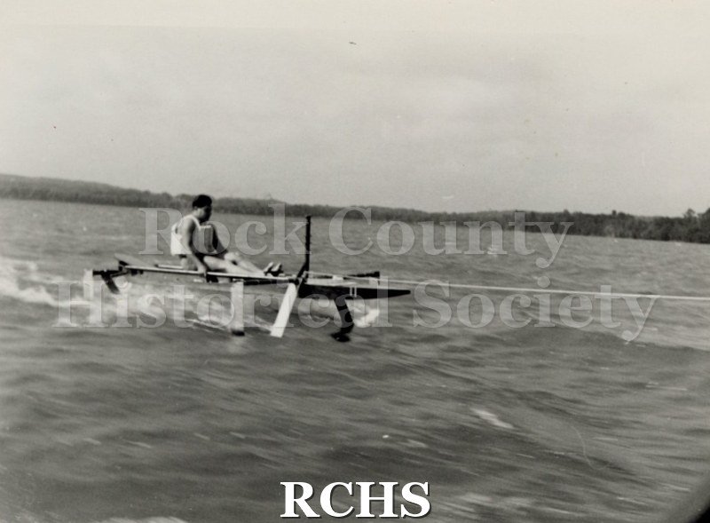 Black and white photograph of Warren Hull sitting in a hydrofoil boat which is moving through the water. There is a shoreline in the background with trees.