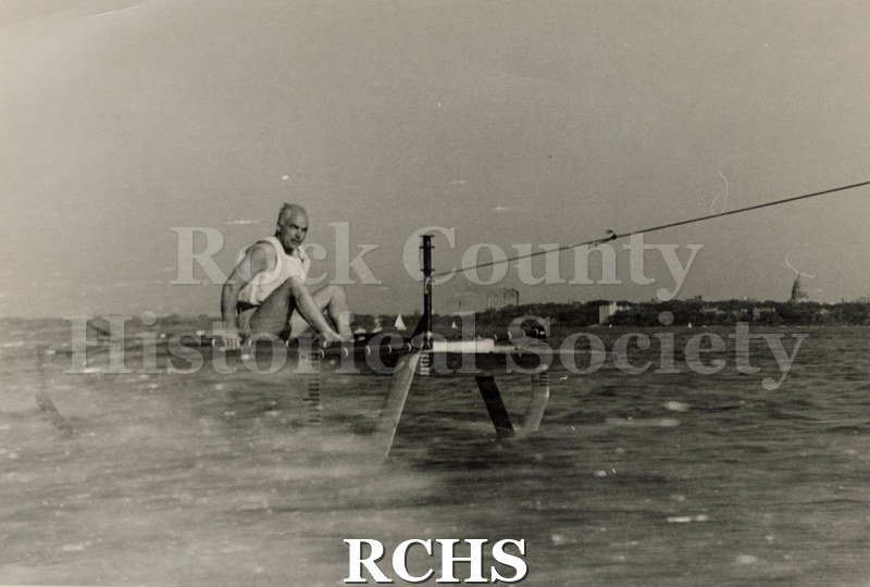 Black and white photograph of J. G.Baker sitting in a hydrofoil boat that is being towed. There is the shoreline of Madison in the background.