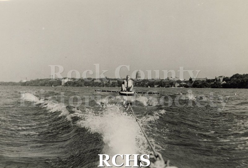Black and white photograph of J. G. Baker sitting in a hydrofoil boat that is being towed. There is the shoreline of Madison in the background.