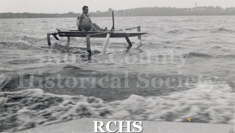 Black and white photograph of Warren Hull sitting in a hydrofoil boat which is moving through the water. There is a shoreline in the background with trees.