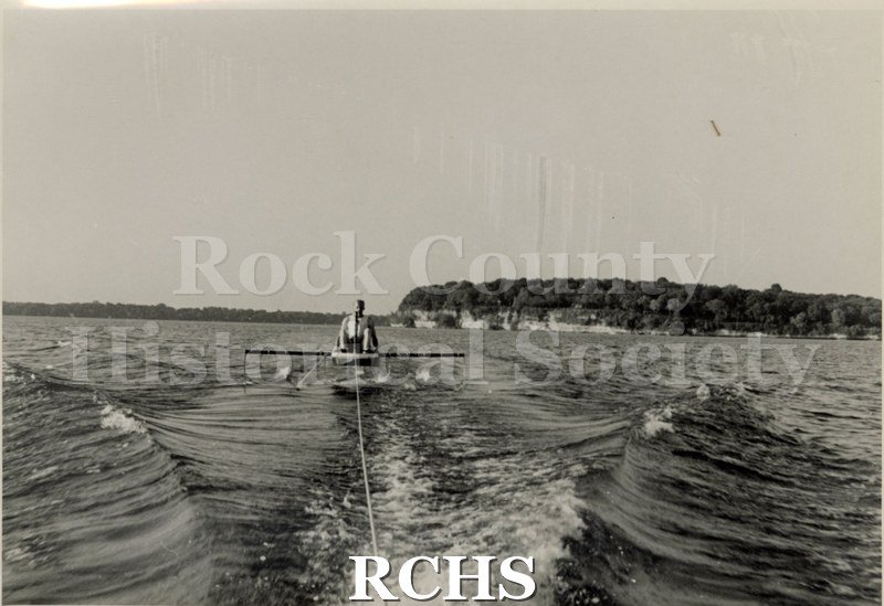 Black and white photograph of J. G. Baker sitting in a hydrofoil boat that is being towed. There is the shoreline of Madison in the background.