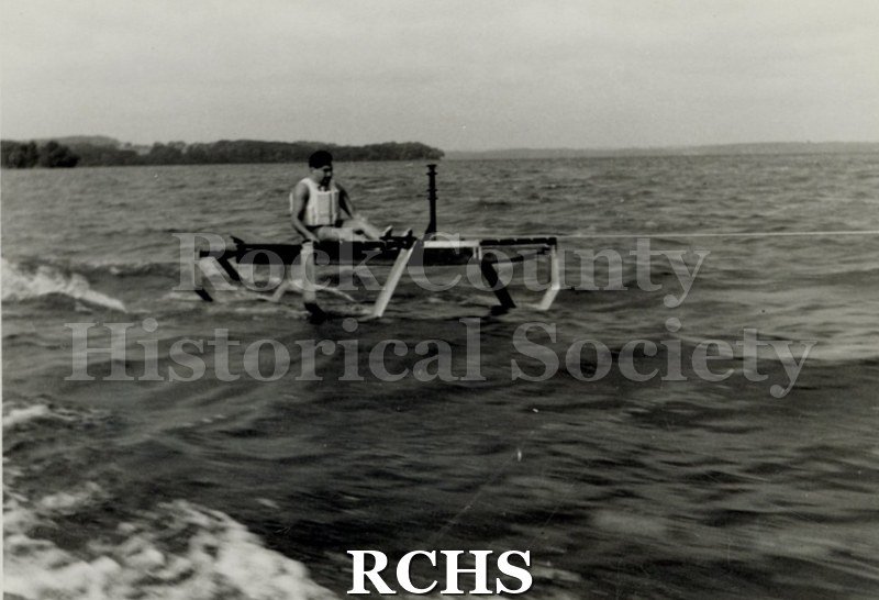 Black and white photograph of Foil testing. Hydrofoil boat is moving through the water with a man sitting it it. There is a shoreline in the background with trees.