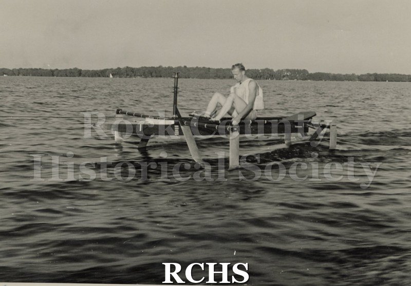 Black and white photograph of Wallace Olsen sitting in a hydrofoil boat that is being towed. Side view of boat and passenger.