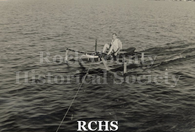 Black and white photograph of Wallace Olsen sitting in a hydrofoil boat that is being towed. Side view of boat and passenger.