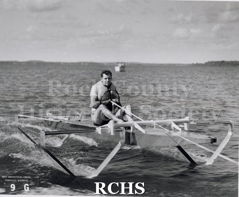 Black and white photograph of a hydrofoil boat with one man aboard, probably Warren Hull. On bottom: "Baker Manufacturing Company Evansville, Wisconsin 9G".
