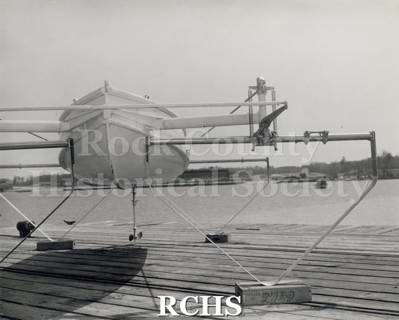 Black and white photograph of a hydrofoil boat on a deck with foils down. One foil is resting on a CocaCola box.