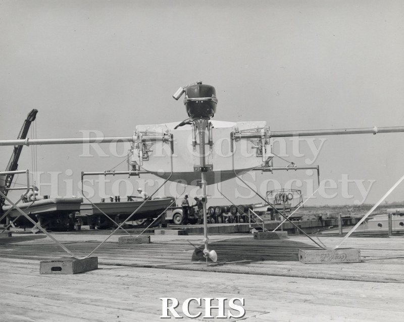 Black and white photograph of a hydrofoil boat on a deck with foils down. Shows back view with engine.