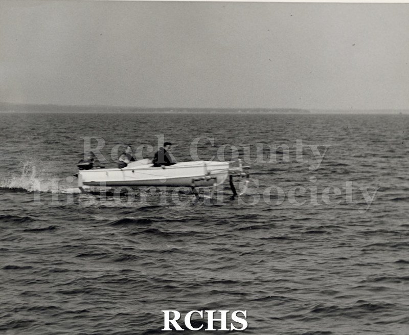 Black and white photograph of a hydrofoil boat on the water with two passengers. Shows a side view.