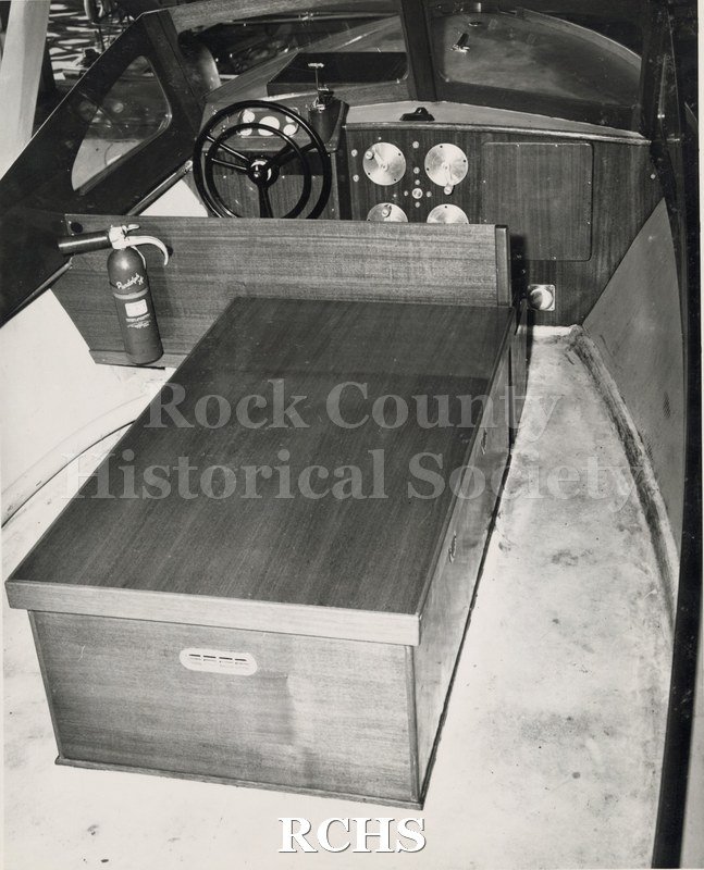 Black and white photograph of a Naval Boat Cockpit. Shows the wheel, dials, fire extinguisher and box.