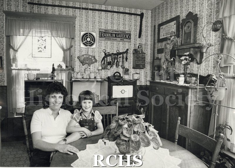 Historic image of 414 E. Holmes, Janesville, WI. Shows the kitchen of a house. There is a young woman and a child sitting at the wooden table.