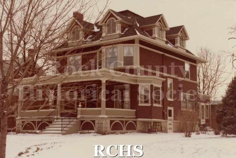 Color photo of 220 Sinclair, Janesville, WI. Shows a three story house with a porch across the front and four dormer windows on the top story.