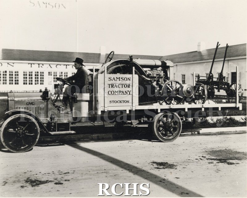 Black and white photograph of a GMC truck with machinery on the bed of the truck and a driver in front. On the side is a sign: "Samson Tractor Company Stockton".