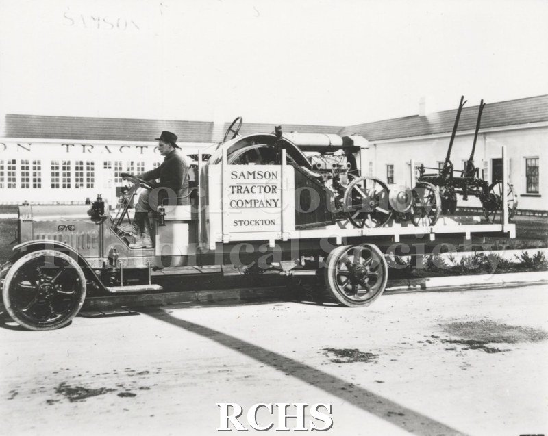 Black and white photograph of a GMC truck with machinery on the bed of the truck and a driver in front. On the side is a sign: "Samson Tractor Company Stockton".