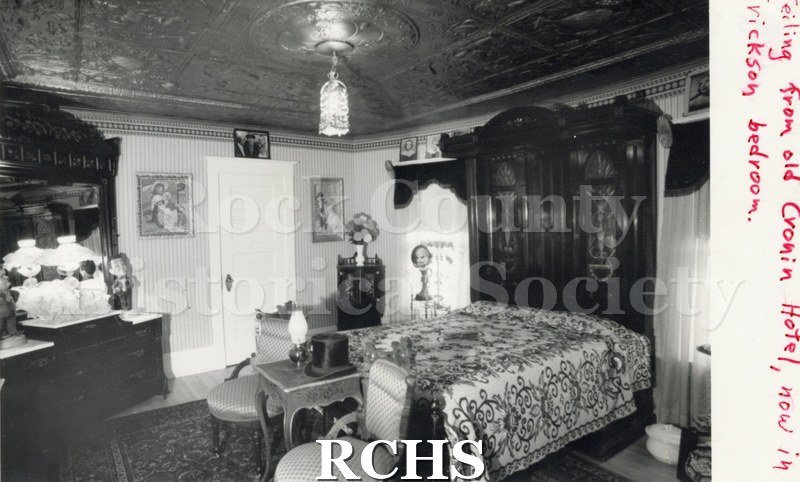 Black and white photograph of the Bennison-Erickson house bedroom with a bed and dresser. Room has an ornate ceiling with a chandelier in the middle.